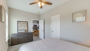 Een slaapkamer met een bed, een dressoir, een plafondventilator en een deur naar een keuken.