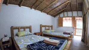A room with two wooden beds, each with a patterned bedspread and pillows. There is a small table with a lamp and a plant on the left side. The room has a wooden ceiling and a window with wooden shutters.