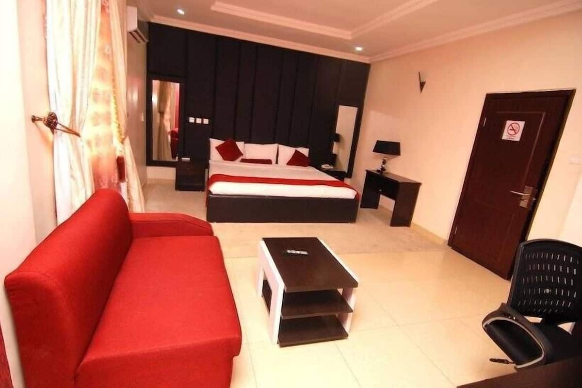 Quarto de hotel com uma cama grande, um sofá vermelho, uma mesinha, uma cadeira e uma porta com o aviso de proibido fumar.
