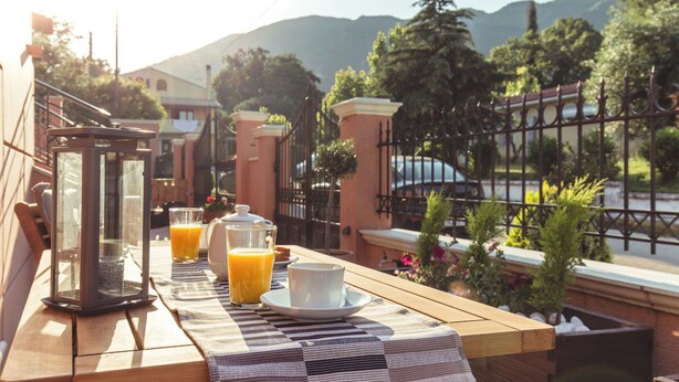 Une table de petit-déjeuner avec du jus d’orange, une tasse et une lanterne, placée sur une terrasse en bois donnant sur un jardin et des montagnes.
