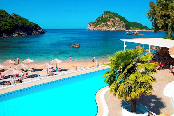 A poolside view with a beach, umbrellas, and a palm tree.
