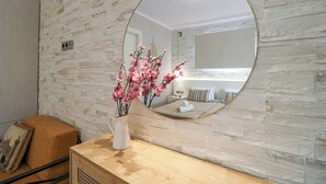 Un baño con pared de piedra, espejo redondo, tocador de madera y un jarrón con flores rosadas.