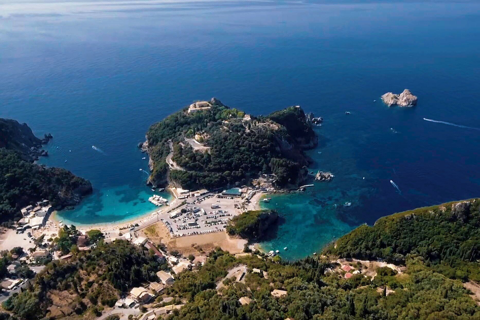 An aerial view of a coastal area with a small island featuring a paved area with buildings, a beach, and clear blue waters.