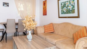 Una sala de estar con un sofá, una mesa de centro y un jarrón con flores secas. Hay sillas, una mesa y un cuadro en la pared.