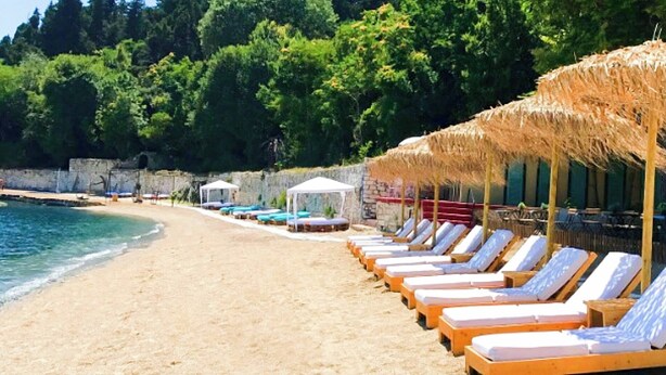 Une plage bordée de rangées de chaises longues et d’ombrelles en chaume, un mur de pierre et une vue dégagée sur la mer.