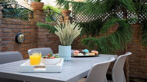 Une terrasse avec une table mise pour le petit-déjeuner, comprenant un plateau avec du jus d’orange et des viennoiseries, entourée de plantes en pot et d’un mur en briques.