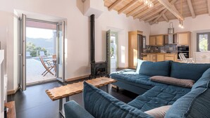 Una sala moderna con un sofá grande, una mesa de centro de madera y un estufa de leña negra. Hay una puerta corrediza de vidrio que da a un patio al aire libre con una mesa y sillas.