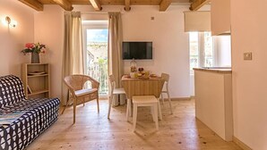 Et hyggeligt værelse med sofa, spisebord og stole. Et fjernsyn er monteret på væggen, og der er et vindue med gardiner.