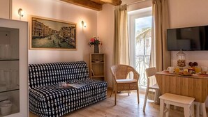 En hyggelig stue med en stribet sofa, en fletstol, et spisebord med morgenmadsprodukter, et fjernsyn og et maleri af et kanalmotiv.