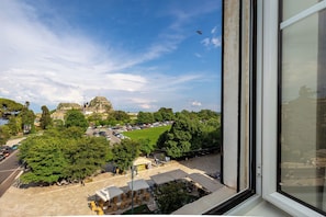 Une vue depuis une fenêtre donnant sur un parc, avec des arbres, une pelouse et un bâtiment situé au loin.