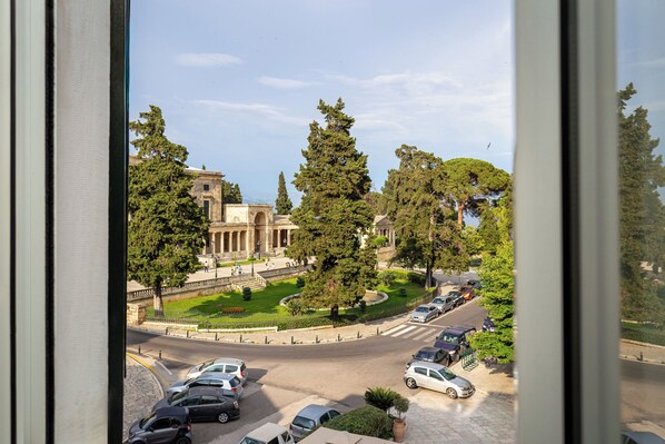 Une vue d’un parking avec des voitures, d’un bâtiment à l’architecture classique et de la verdure environnante.