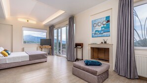 Ein modernes Schlafzimmer mit Bett, einer Couch, einem Fernseher, einem Kamin und einem großen Fenster mit Vorhängen.
