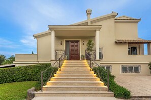 Un bâtiment de couleur beige, doté d’une entrée centrale, d’un escalier et d’une zone pavée.