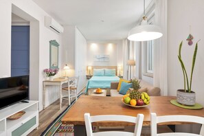 A modern hotel room with a wooden dining table, white chairs, a flat-screen TV, a bed with blue bedding, and a large window.