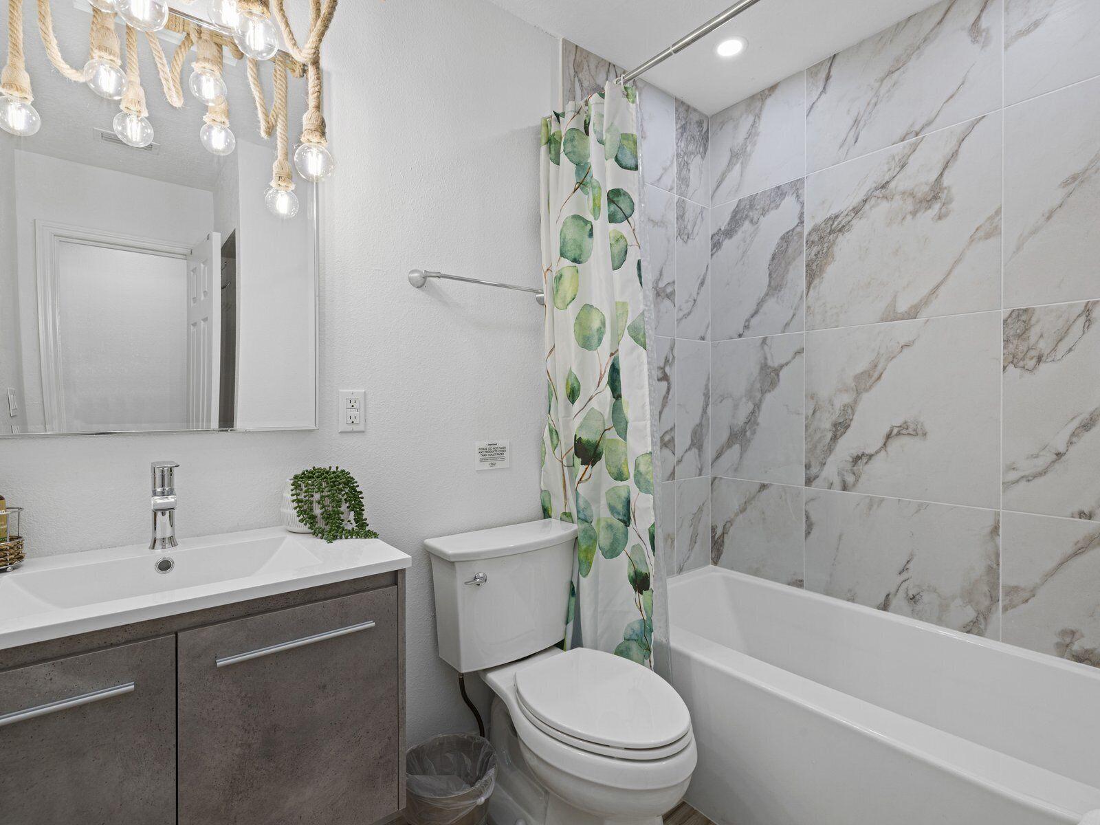 Un baño con pared de ducha de mármol, inodoro, lavamanos con espejo y candelabro con bombillas transparentes.
