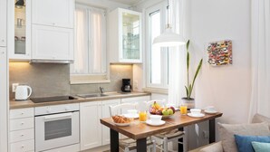 A modern kitchen with white cabinetry, a built-in oven, a wooden dining table, and a breakfast setup with fruits and a coffee maker. - Studio No2, CasaNova, Corfu old town center (Corfu)