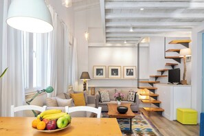 A modern living room with a sofa, coffee table, and a fruit bowl. There is a staircase with a TV on the upper landing.
