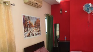 A room with a bed, two sofas, a glass-top coffee table, an air conditioner, a painting, a mirror, and a red wall. - Lilawati's Blue Fox (Sonapur)