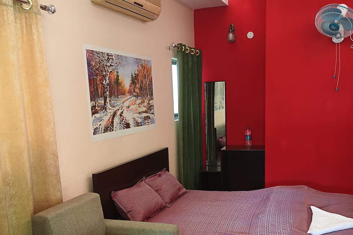 A room with a bed, two sofas, a glass-top coffee table, an air conditioner, a painting, a mirror, and a red wall.
