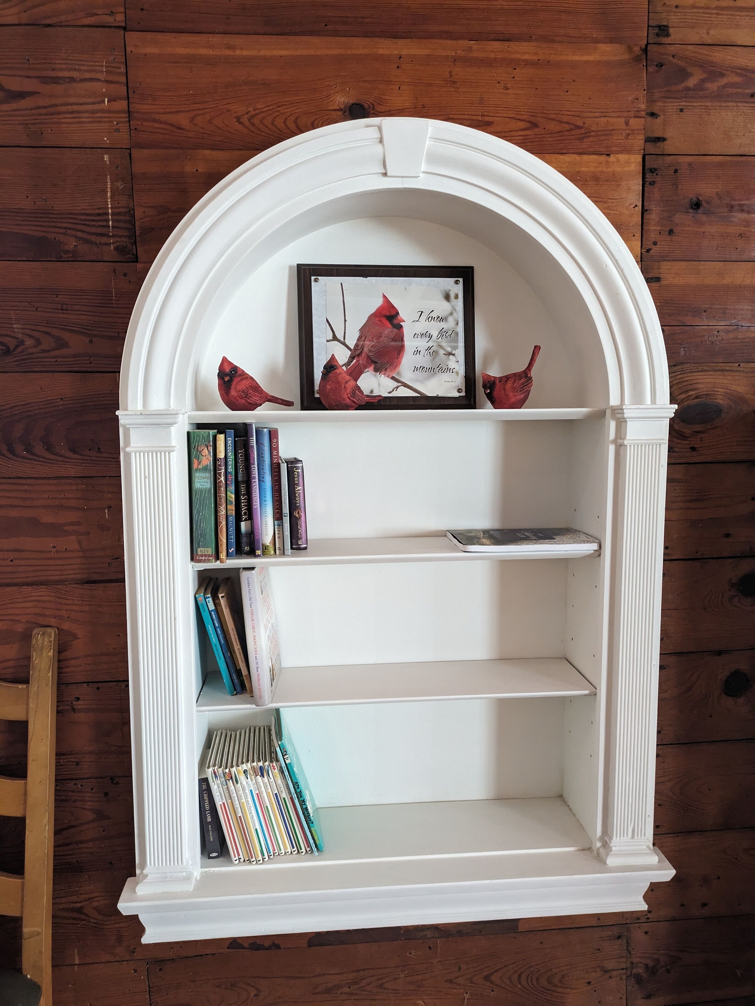 A white, arched bookshelf with books and a framed picture of cardinals.