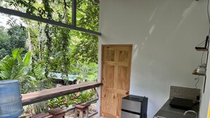 Une cuisine compacte équipée d’un réfrigérateur, d’un évier et d’une porte en bois. Un petit coin repas, avec une table et des tabourets, est situé à côté de la cuisine et offre une vue sur un jardin luxuriant.