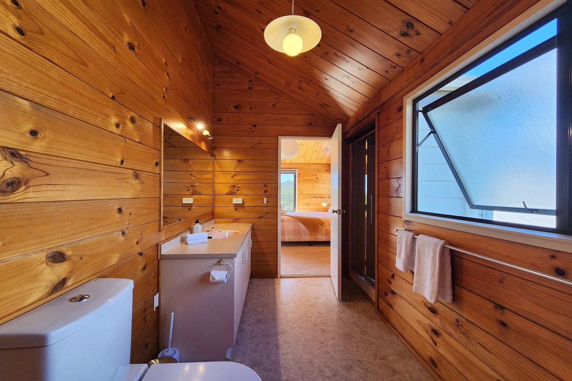 A wooden cabin interior with a bathroom, a bed, and a large window.