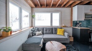 A cozy living room with a gray sectional sofa, a wooden ceiling, and large windows.