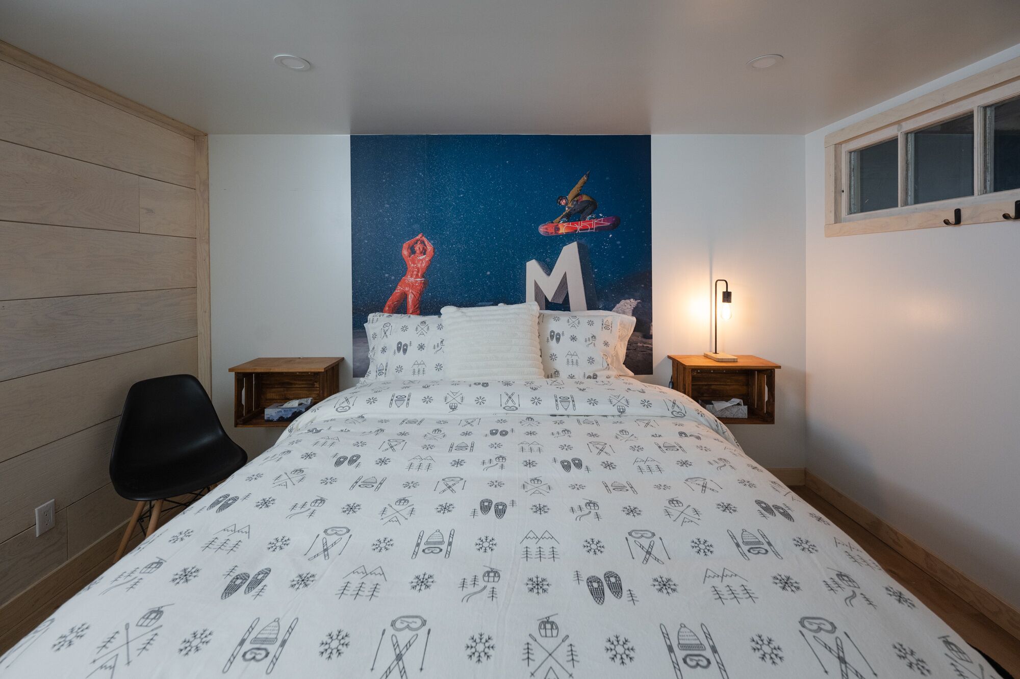 Un dormitorio con un mural de noche estrellada, una cama con sábana blanca que presenta iconos de esquí y snowboard, una silla negra y dos mesitas de noche con lámparas.