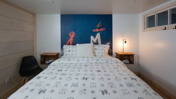 Un dormitorio con un mural de noche estrellada, una cama con sábana blanca que presenta iconos de esquí y snowboard, una silla negra y dos mesitas de noche con lámparas.
