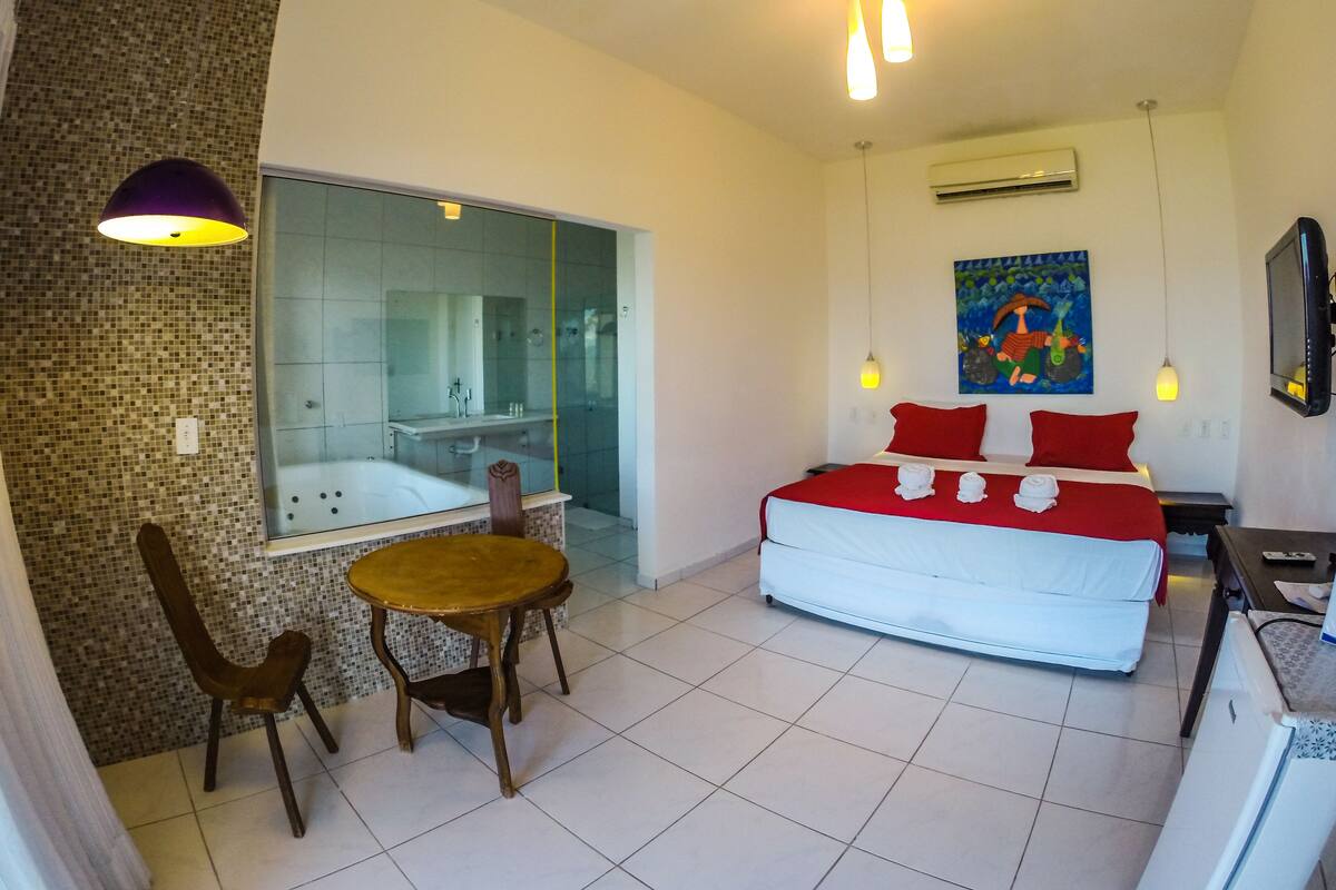 A modern hotel room with a large bed, a small table, a chair, a glass-enclosed shower, and a TV.