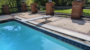 Una piscina rectangular con borde de azulejos, rodeada por un muro de ladrillo y una valla metálica.
