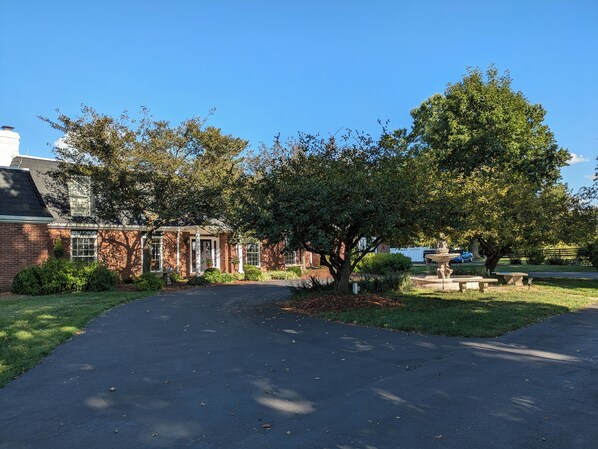 A brick house with a paved driveway, a large tree, and a fountain in the yard.