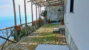 A covered outdoor area with a stone pathway leading to a white wall, a wooden pergola, and a view of the sea.