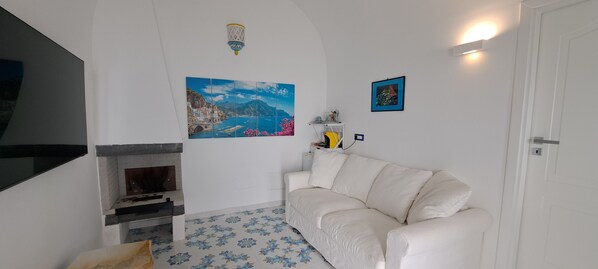 A living room with a white sofa, a fireplace, a painting of a coastal scene, and a small shelf with decorative items.