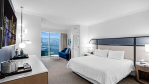 A hotel room with a large bed, a desk with a lamp, a chair, and a view of the city through the window. - Junior Suite Ocean View Floor Fontainebleau (Miami Beach)