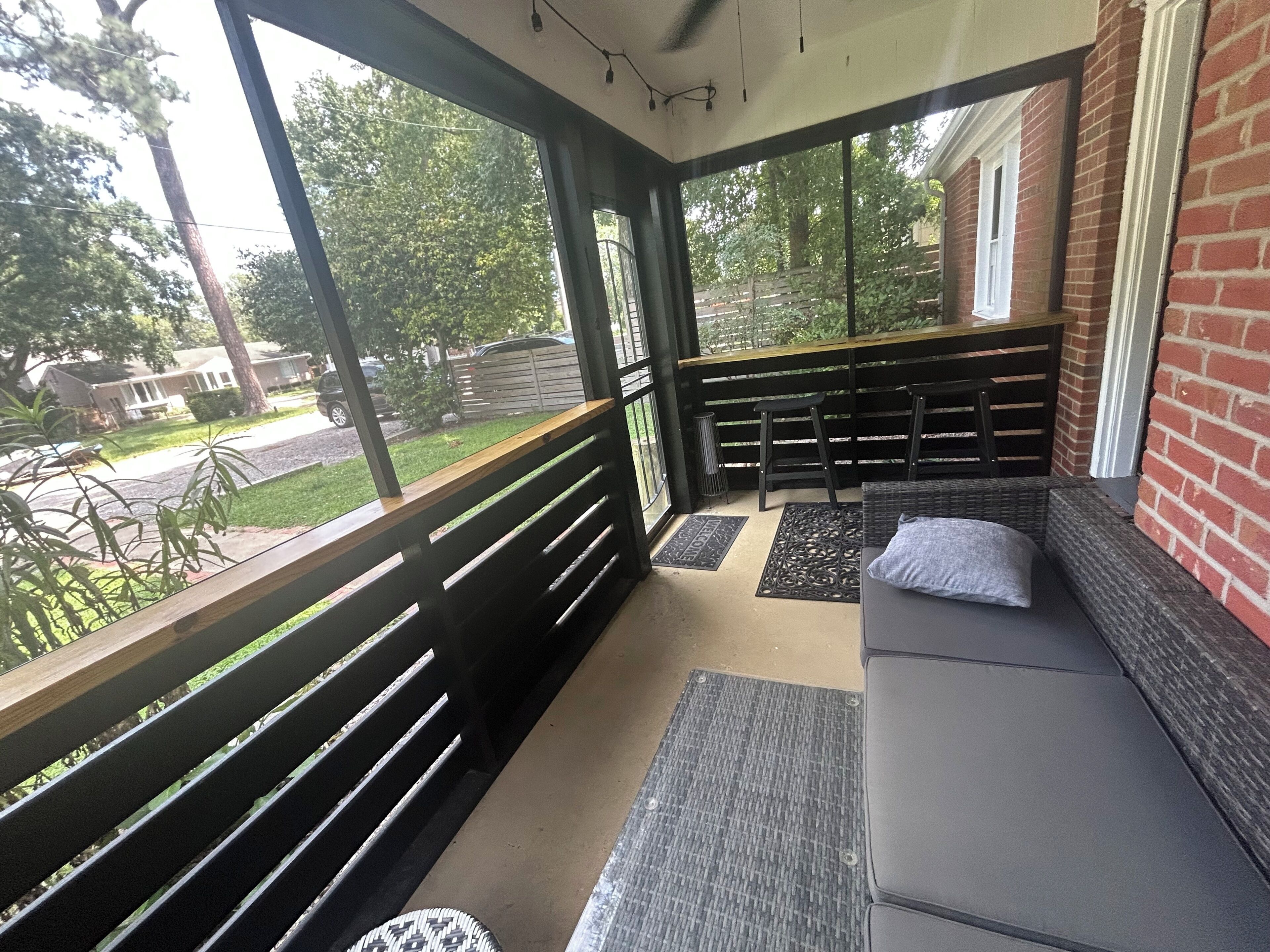 A covered outdoor seating area with a bench and a cushioned bench, a doormat, and a view of the street and greenery.