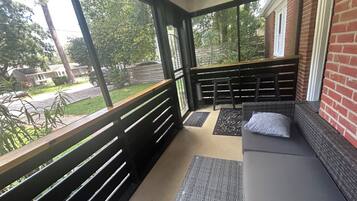 A covered outdoor seating area with a bench and a cushioned bench, a doormat, and a view of the street and greenery.