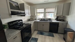 A compact kitchen with a stove, oven, sink, and dishwasher. There are cabinets, a window with blinds, and a small trash bin. - 6 Block to oceanfront and in the center of the vibe district! (Virginia Beach)