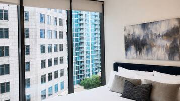 A modern hotel room with a large bed, a view of a high-rise building, and a painting on the wall.