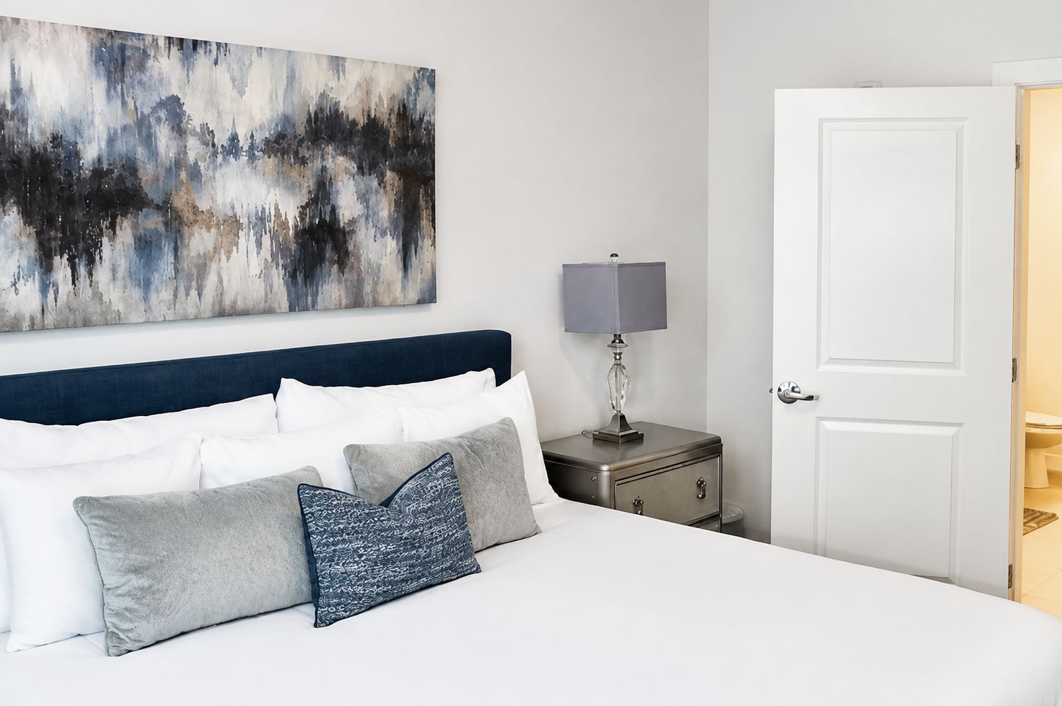 Habitación de hotel con cama, mesita de noche con lámpara, una puerta y un cuadro abstracto grande en la pared.