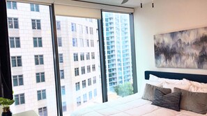 A bedroom with a large window, a bed with pillows, and a painting on the wall.