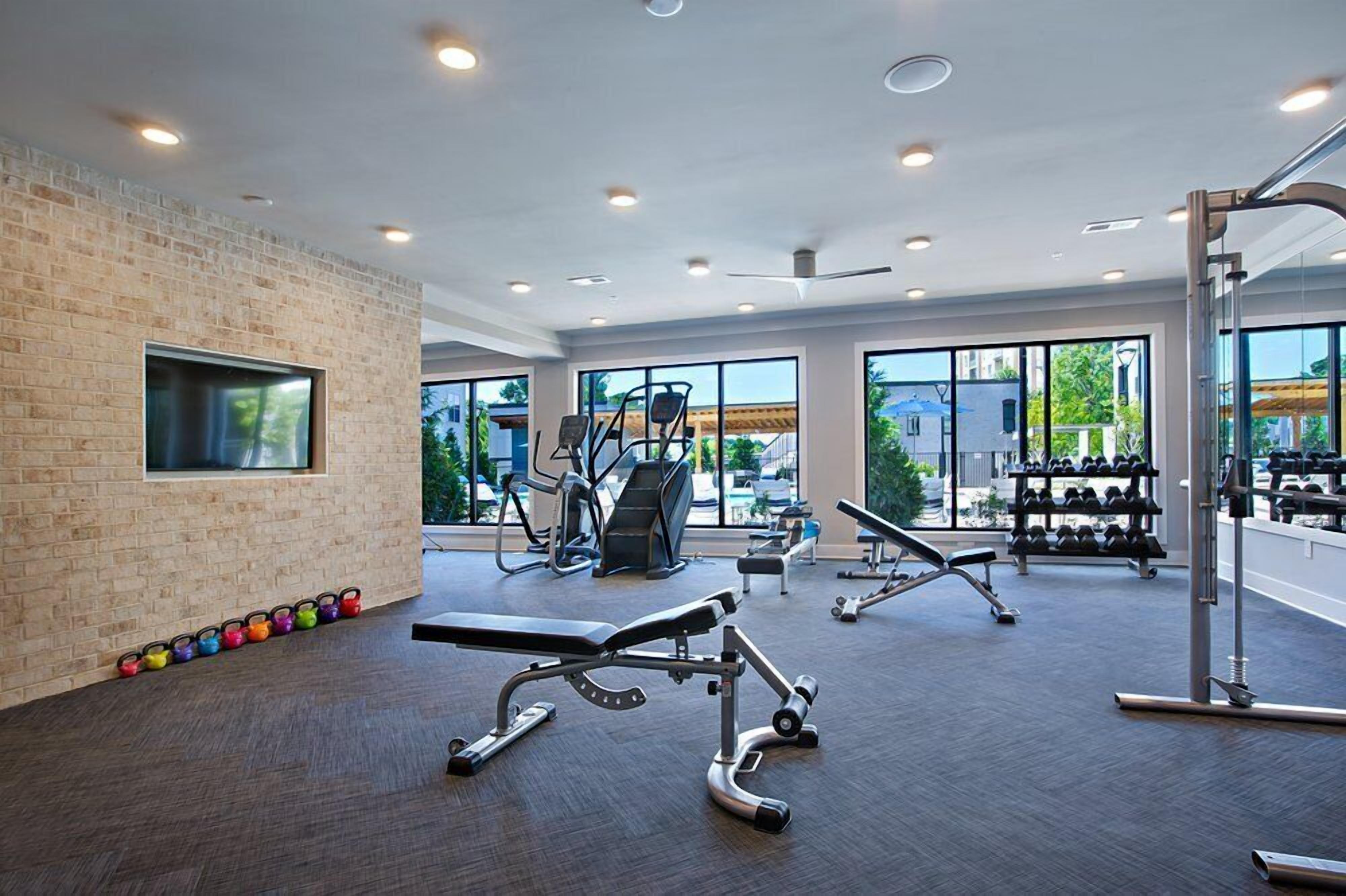 Un gimnasio moderno con diversos equipos de ejercicio, ventanas amplias y una pared de ladrillo.