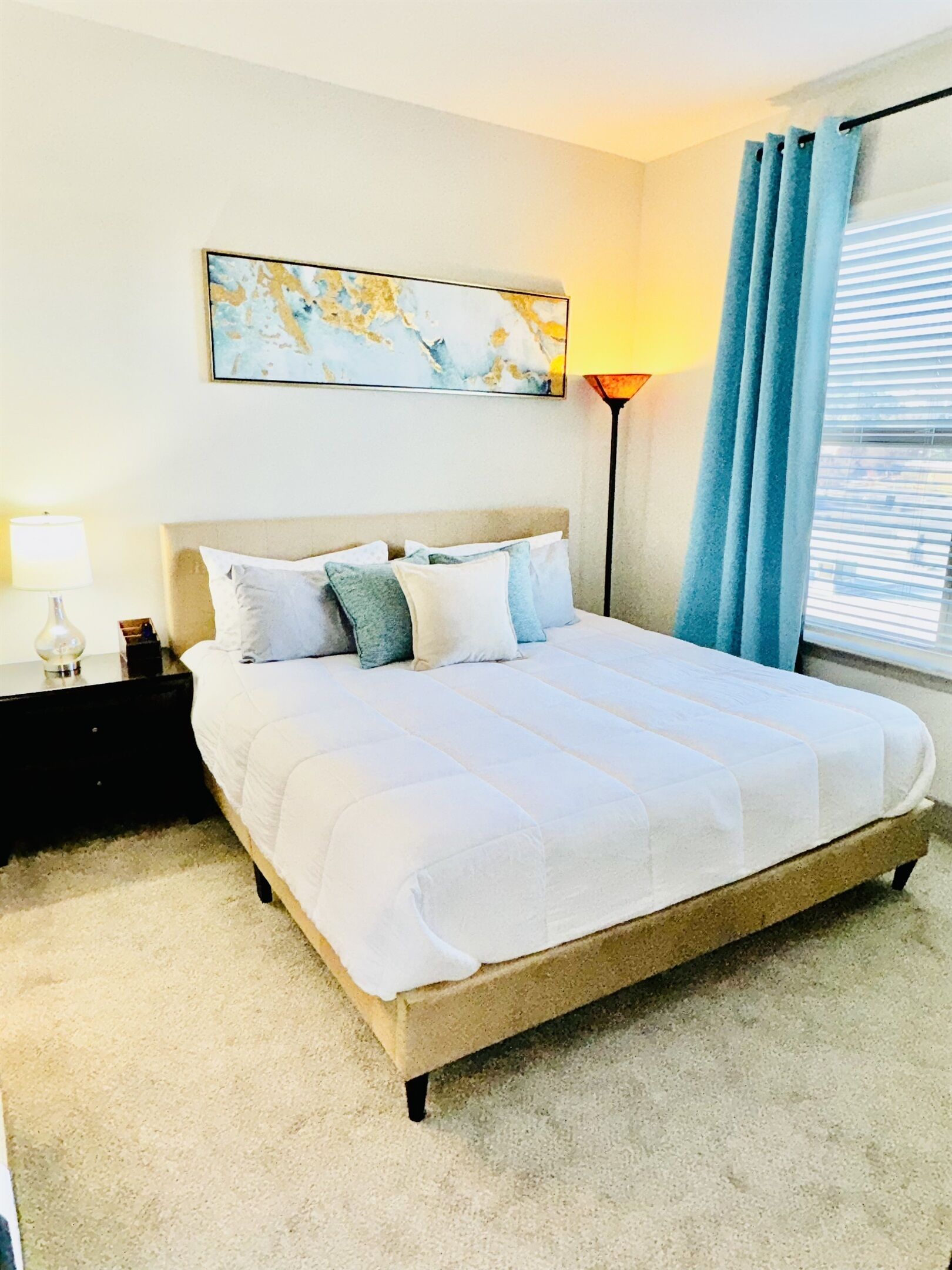 A neatly made bed with a white comforter and multiple pillows, a bedside table with a lamp, a wall-mounted abstract painting, and a window with blue curtains.