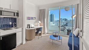 A modern hotel room with a kitchenette, a dining area, a sofa, and a large balcony with a city view.