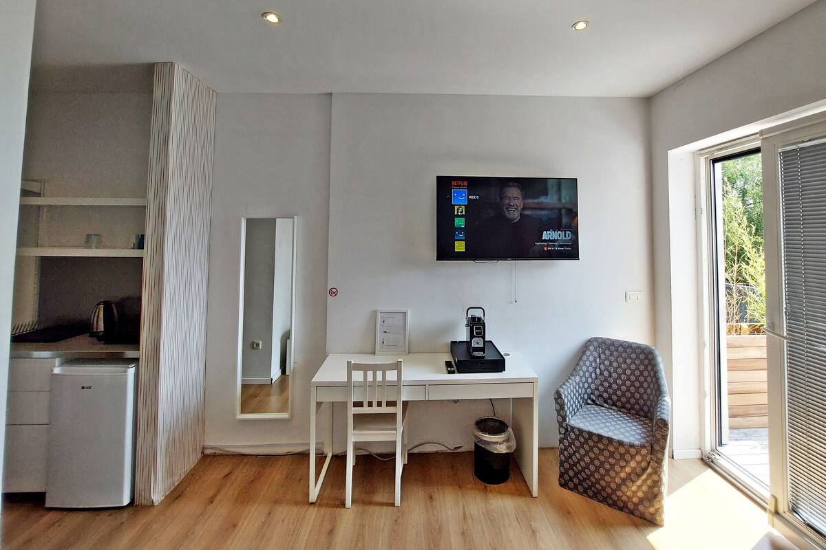A modern room with a flat-screen TV mounted on the wall, a white desk with a chair, a small kitchenette, and a large window with blinds.