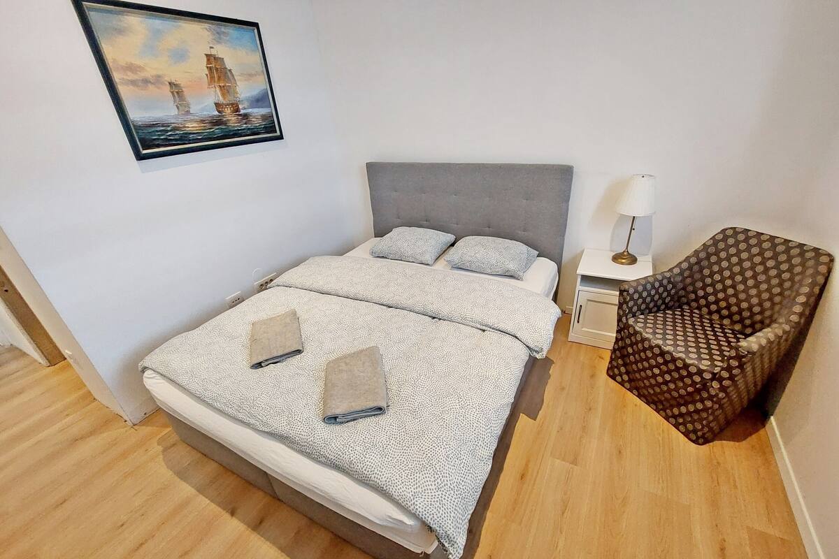 A neatly arranged bedroom with a large bed, a side table with a lamp, a polka-dotted chair, and a framed painting on the wall.