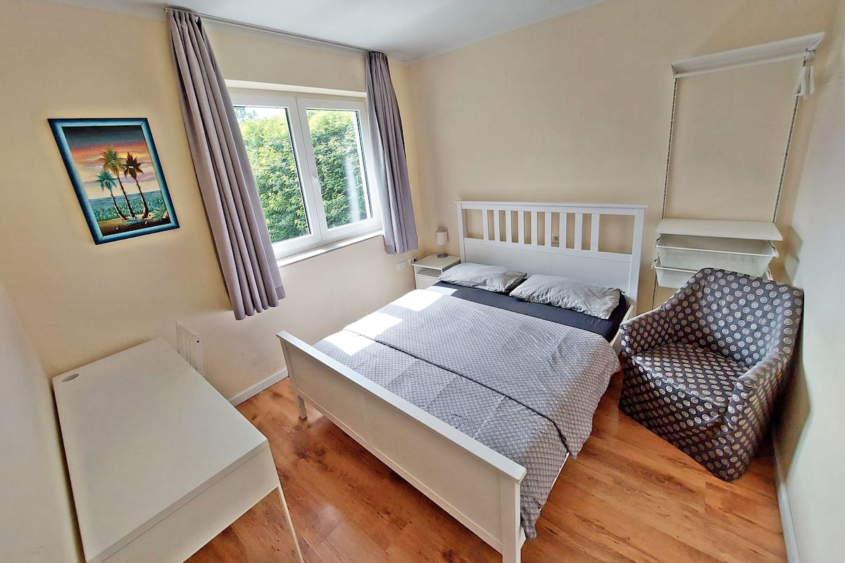 A bedroom with a bed, a chair, a desk, a window with curtains, and a painting on the wall.