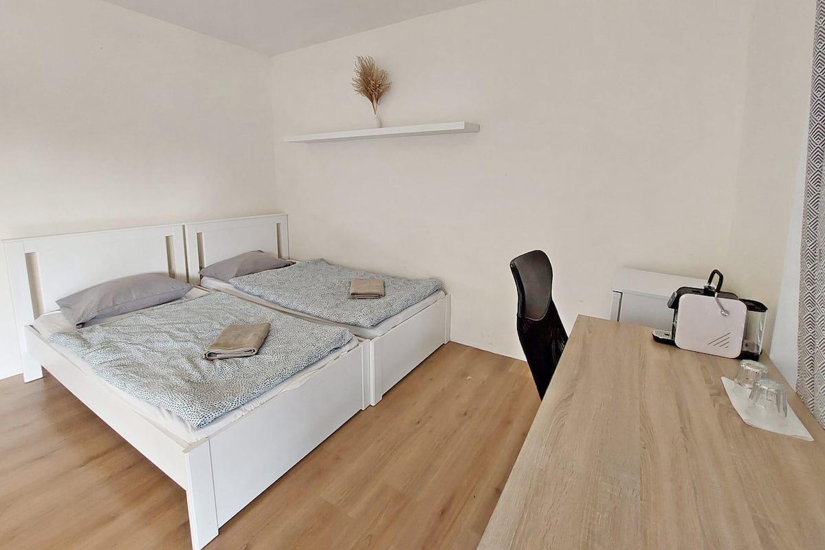 A small, clean room with a bed, a chair, a table, and a shelf with a decorative item.