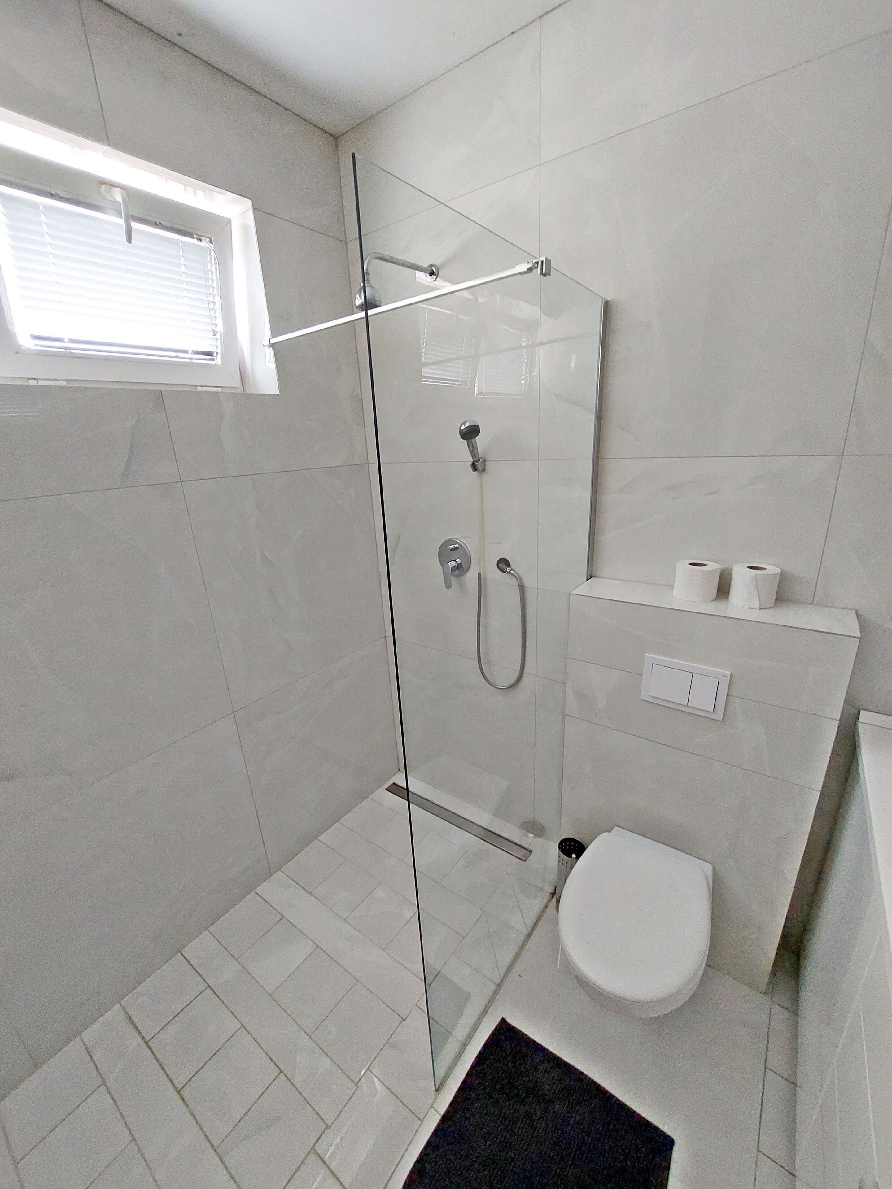 A modern bathroom with a glass-enclosed shower, a toilet, and a small window.