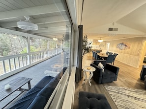 A modern living room with a large window, a dining area, and a kitchen visible through the glass wall.